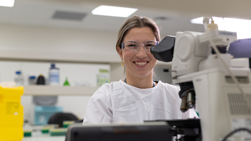 A smiling woman in a lab coat examines a microscope, showcasing her enthusiasm for scientific research.