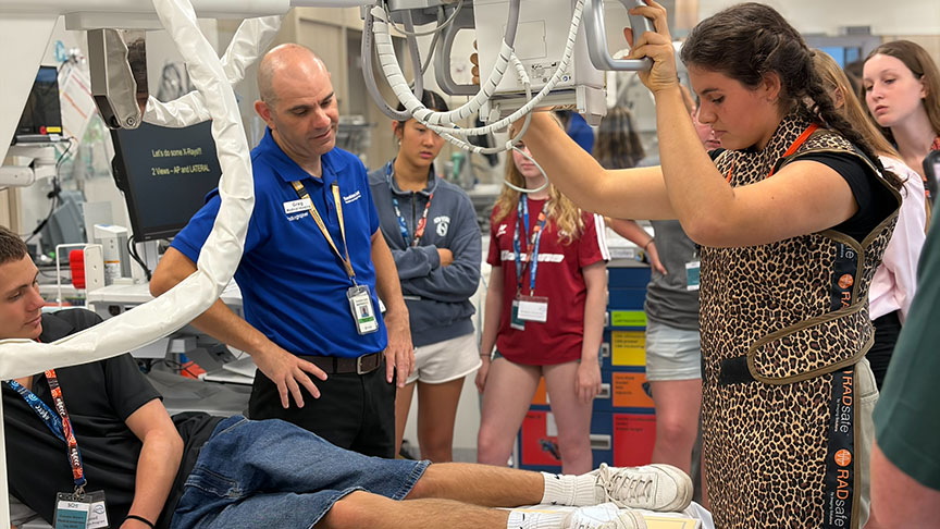 In a hospital setting, a woman supports a man's leg, indicating a medical procedure or assistance.