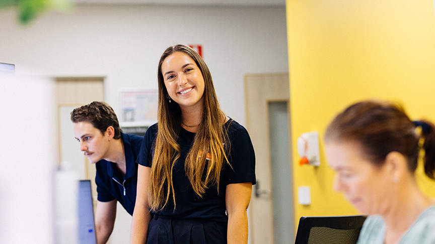 A young woman smiles while standing in front of a computer, exuding confidence and positivity in a professional setting.
