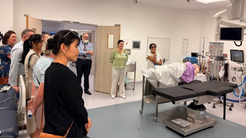 A group of people stands around a hospital bed, attending to a patient in a well-lit hospital room.