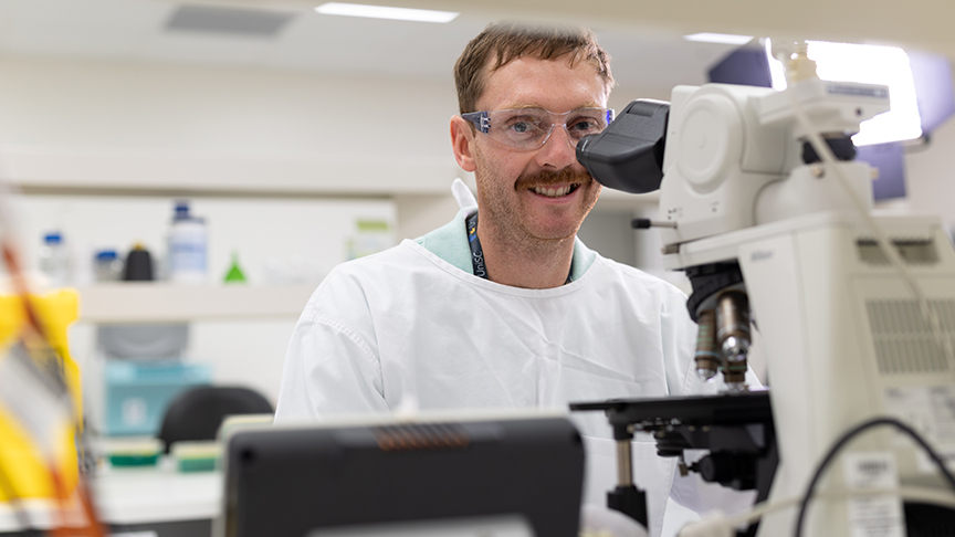 A man in a lab coat examines a specimen through a microscope in a laboratory setting.