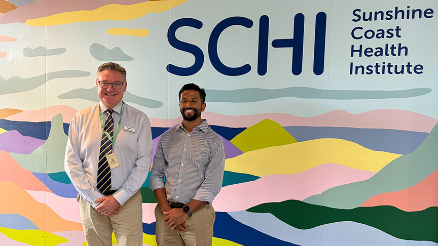 Two men pose in front of a wall featuring the Sunshine Coast Health Institute logo, with bright sunlight illuminating the scene.