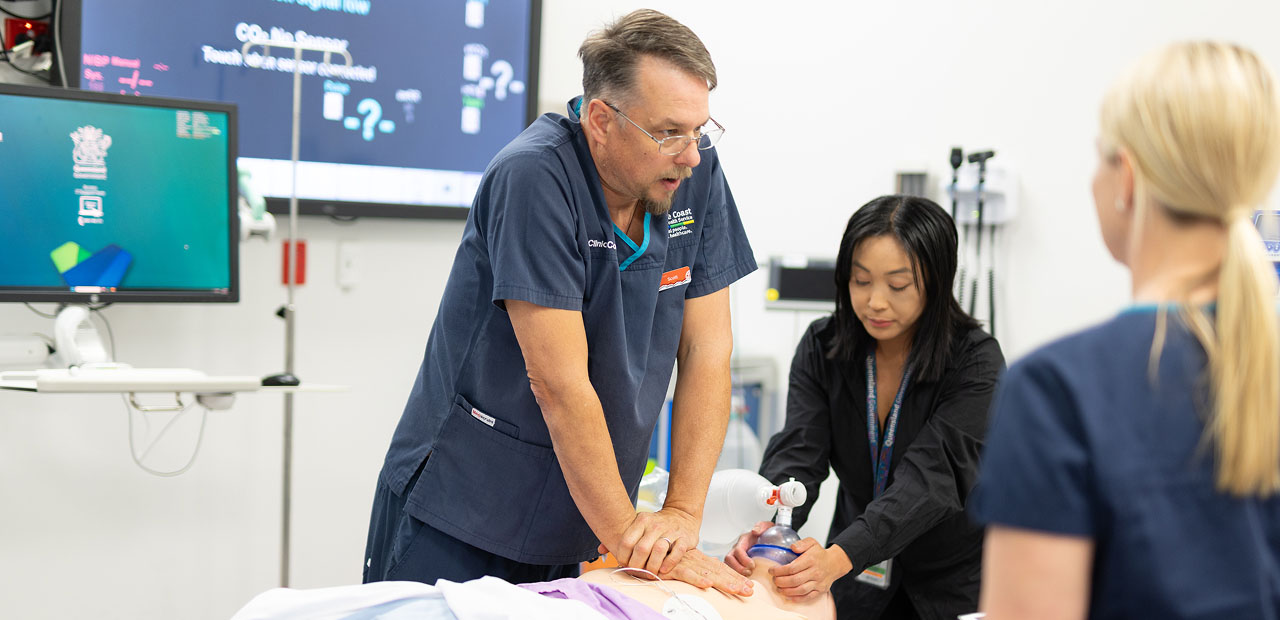 A man is performing chest compressions on a maniki while one woman performs a bag-valve-mask ventilation on the maniki. Another woman stands beside to observe.