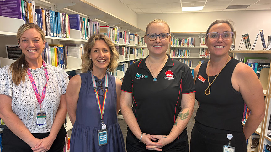 Four women stand in front of tall bookshelves filled with books in a well-lit library setting.